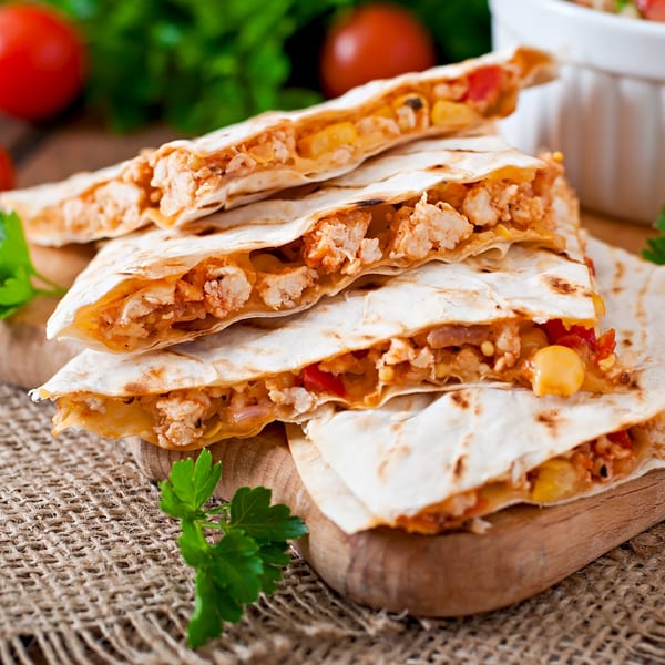 Stacked chicken quesadillas with corn and peppers on a wooden board, with tomatoes and parsley in the background.