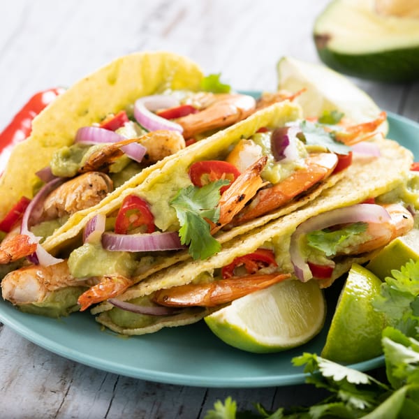Three shrimp tacos with avocado, red onion, chili, and lime wedges on a blue plate.