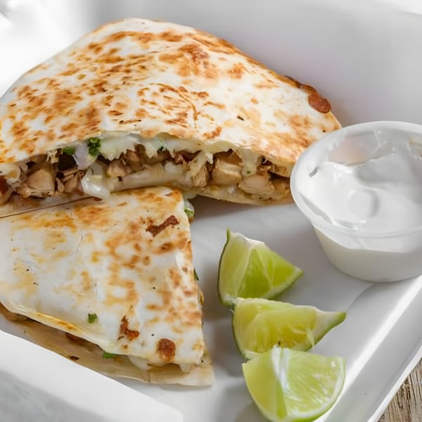 Chicken quesadilla from Los Comparres, cut in half, with lime wedges and sour cream on a white plate.
