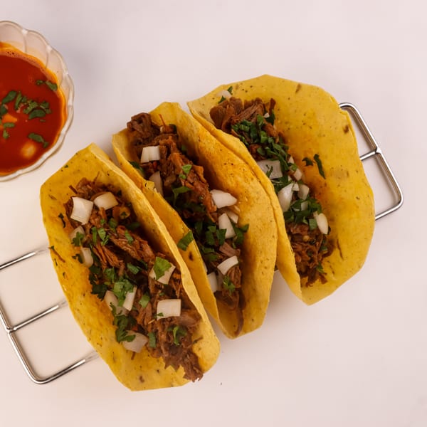 Three Los Comparres birria tacos with meat, onions, and cilantro, with a side of consommé.